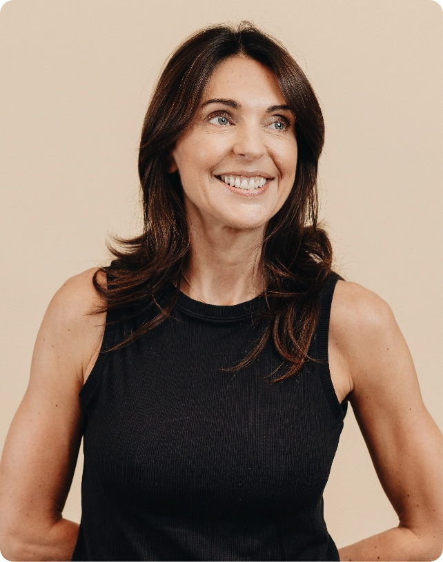 Smiling woman with long brown hair in a black sleeveless top against a beige background.