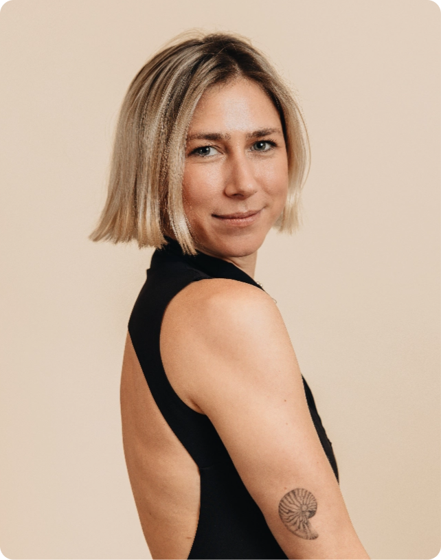 Woman with short blonde hair in a black sleeveless top, showing a tattoo on her upper arm, smiles slightly against a beige background.