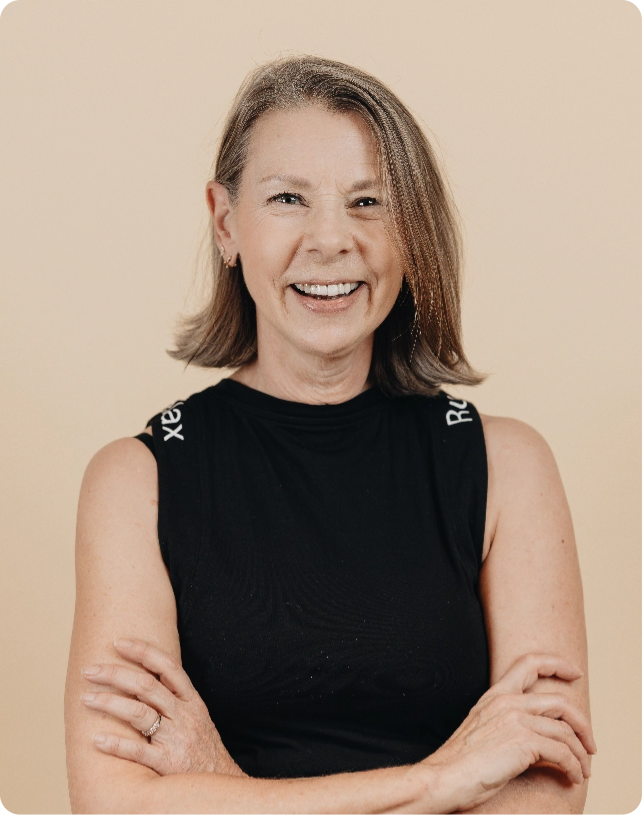 Woman with shoulder-length hair in a black sleeveless top stands smiling with arms crossed against a beige background.