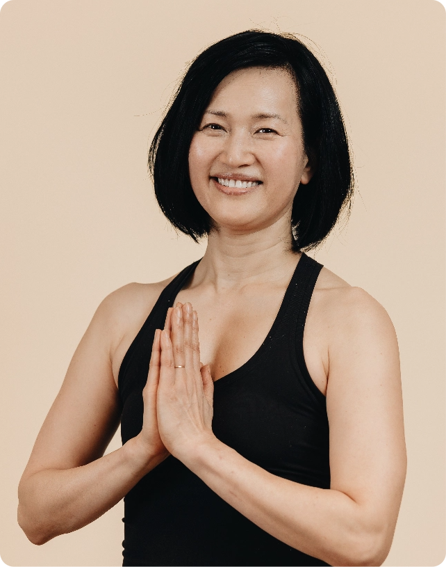 Smiling woman in a black tank top with short dark hair, standing with hands in prayer position against a neutral background.