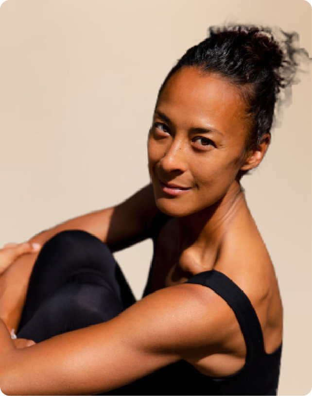 Person sitting with knees up, wearing a black tank top, looking at the camera with a gentle smile against a plain background.