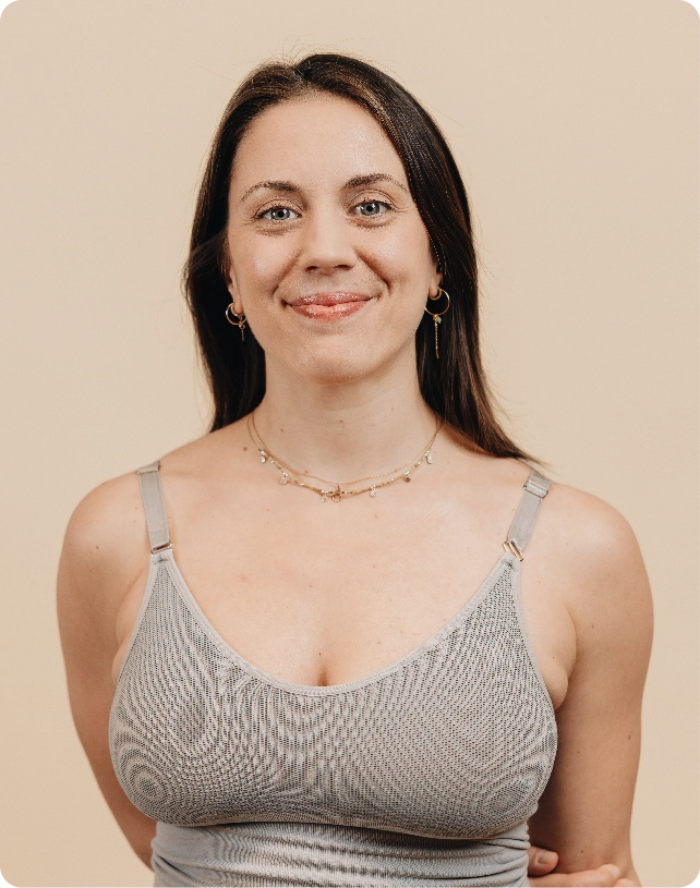 A woman with long dark hair wearing a gray tank top and a delicate necklace, smiling against a beige background.