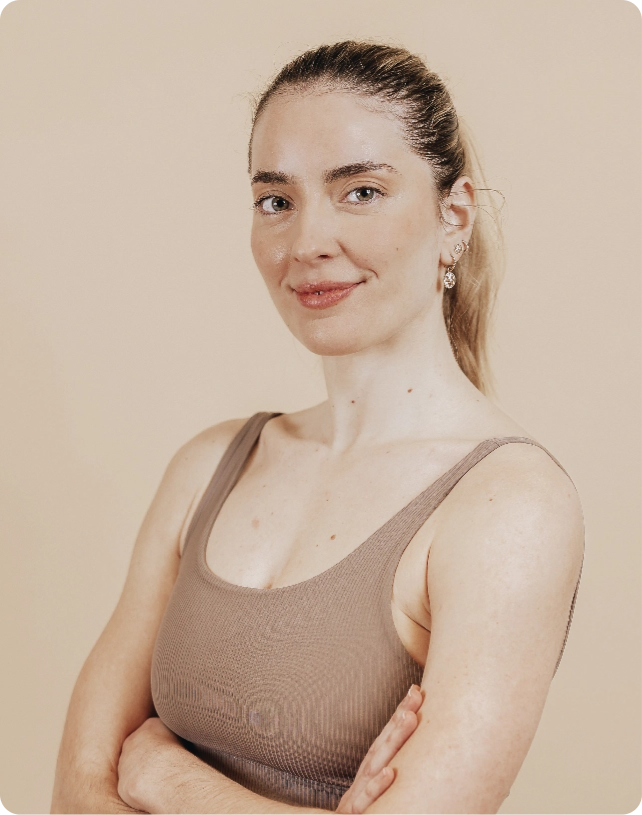 Smiling woman with light brown hair in a ponytail, wearing a beige tank top, standing with arms crossed against a beige background.