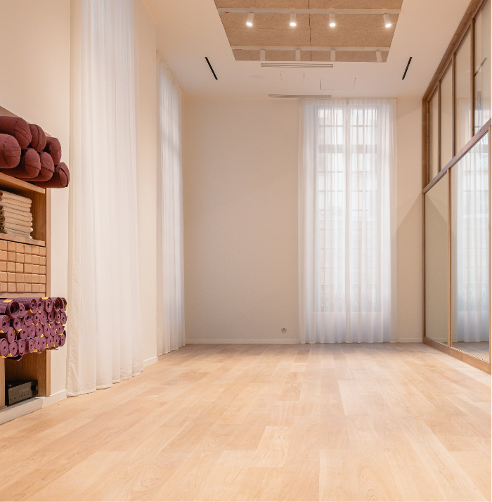 Bright, empty yoga studio with wooden floors, stacked mats and towels, large mirrors, and sheer curtains filtering natural light.