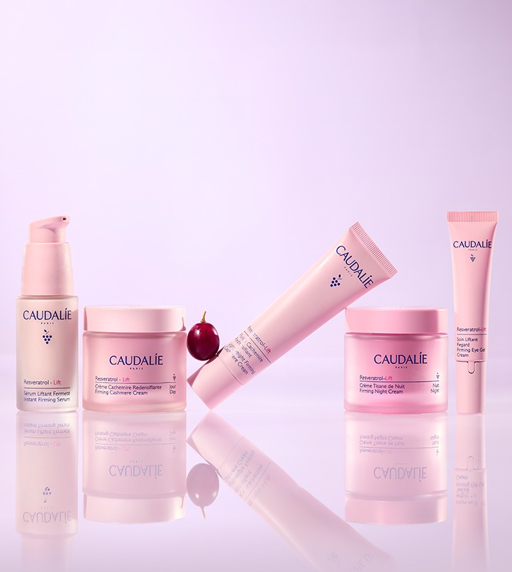 Caudalie skincare product collection arranged on lavender background with pink bottles and jars, featuring serums, creams, and tubes alongside a red plum.
