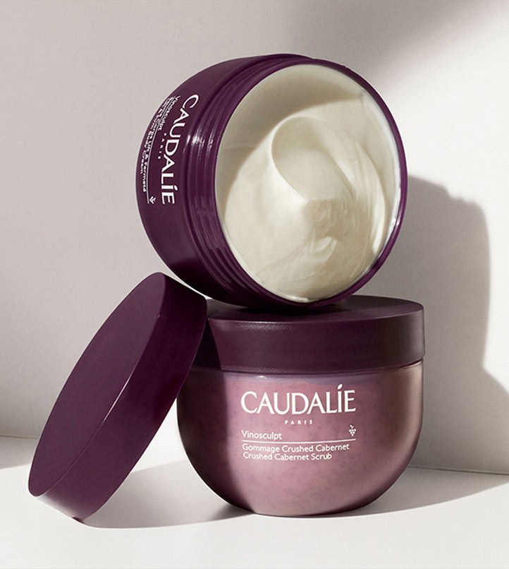 Two open purple jars of Caudalie Vinosculpt Crushed Cabernet Scrub, one on its side with creamy scrub visible, against a light background.