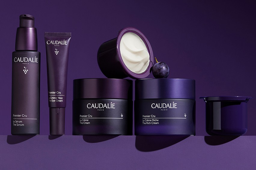 Caudalie skincare products on a purple background, including serum, eye cream, and rich cream, with a grape accent.